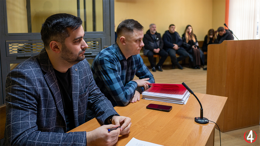 На фотографії зображено сцену у залі суду. На передньому плані за столом сидять двоє чоловіків. Ліворуч чоловік у темному піджаку в клітинку, праворуч – чоловік у картатій сорочці. На столі перед ними лежать стоси документів, червона папка, смартфон і мікрофон. На задньому плані сидять інші люди, ймовірно, учасники або глядачі судового процесу. Приміщення має жовті стіни, дерев'яну підлогу та кафедру для виступів у правій частині кадру